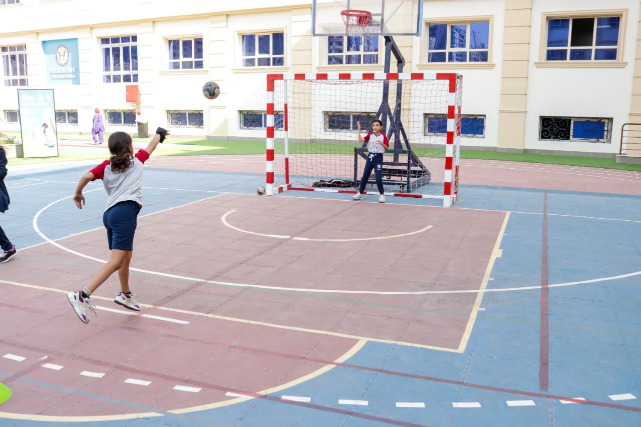 This image showcases an exciting sports activity taking place on the outdoor court at IVY STEM International School. A girl is seen throwing a ball towards a goal, while another girl stands ready to block or catch the ball. The court is designed for multiple sports, including handball and basketball, as indicated by the colored lines and hoops. The school setting is evident from the architecture of the buildings in the background and the school emblem on the wall. A teacher or supervisor can be seen overseeing the activity, adding to the vibrant atmosphere. The presence of a soccer ball near the goal highlights the multi-sport functionality of the court.